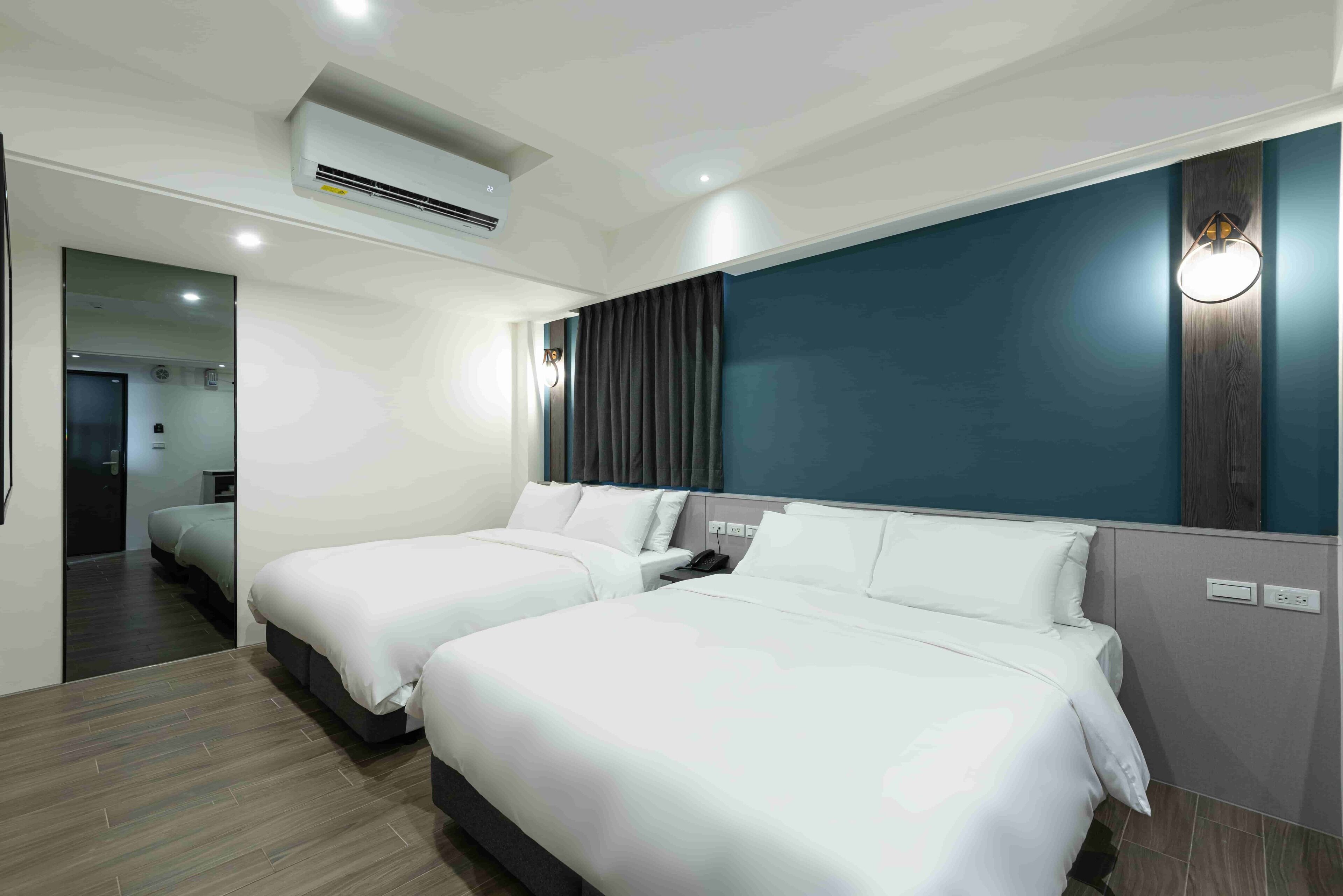 A hotel room with two beds, a large mirror, an air conditioning unit, a dark-colored wall, and a light fixture.