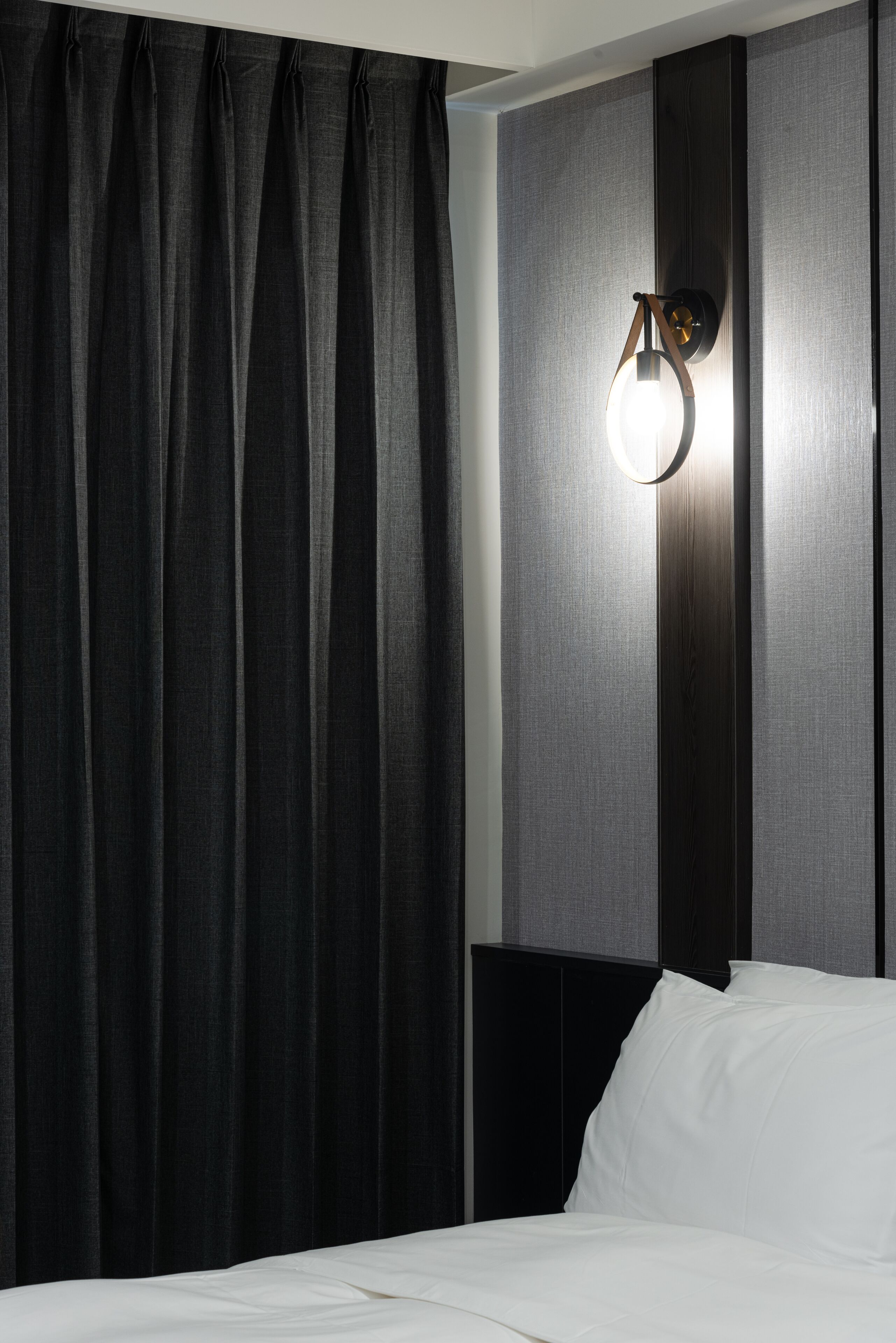 A hotel room with a bed, black curtains, a wall-mounted light fixture, and a textured wall panel.