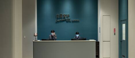 Un mostrador de recepción de hotel con una pared azul oscuro y un letrero que dice "Hotel Euphemia". Se ven dos recepcionistas detrás del mostrador, y una puerta con un letrero rojo de salida de emergencia a la derecha.