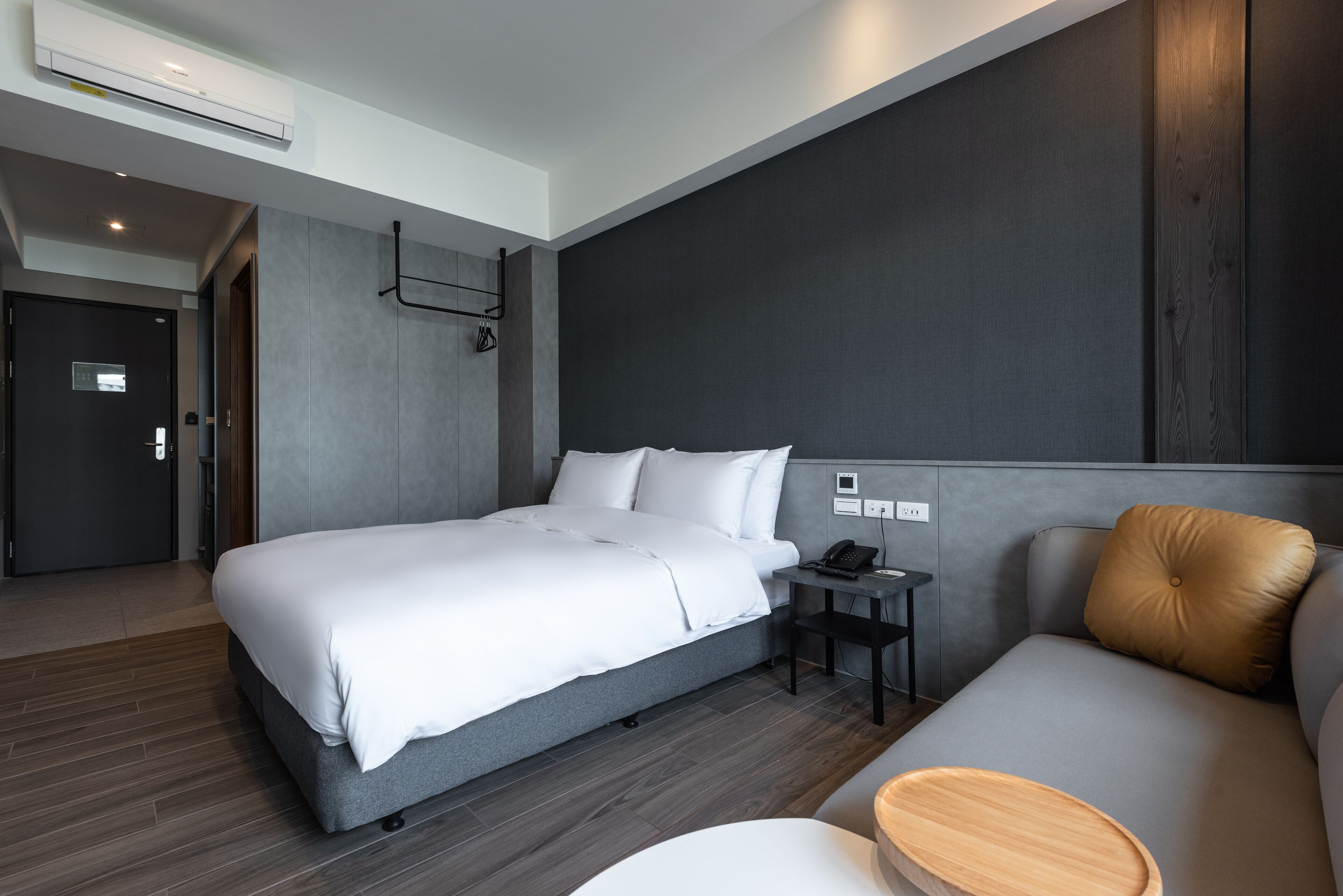 A modern hotel room with a large bed, a bedside table with a phone, a small table with a bowl, and a sofa with a cushion.