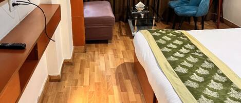 Una habitación de hotel con suelo de madera, una cama con edredón verde y blanco, un televisor montado en la pared, una mesita con lámpara y una silla azul.