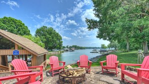 A lakeside deck with red Adirondack chairs, a fire pit, and a view of a dock and lake. - Stay Awhile by AvantStay | Lakefront + Pool, Dock (Mooresville)