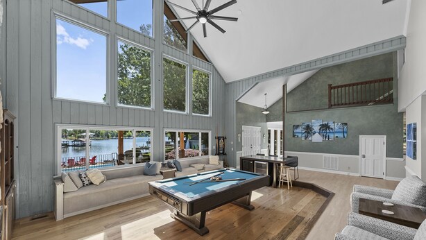 A spacious living room with a pool table, large windows overlooking a body of water, and a high ceiling with a fan. - Stay Awhile by AvantStay | Lakefront + Pool, Dock (Mooresville)
