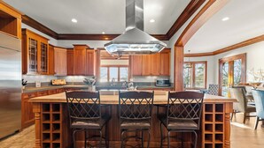 A kitchen with wooden cabinets, a stainless steel refrigerator, a central island with bar stools, and a large overhead vent.