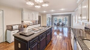 A spacious kitchen with dark cabinetry, a granite countertop, and a central island. There is a dining area with a table and chairs visible through the open door. - Lakefront Home w/ Prv. Dock, Tiki Bar & Firepit | Shore to Please by AvantStay (Denver)