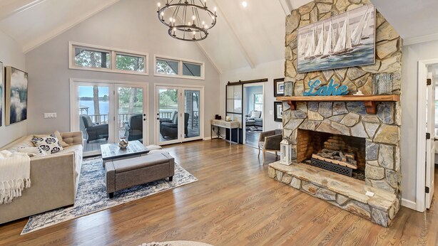A spacious living room with a stone fireplace, large windows, and a chandelier. - Lakefront Retreat • Hot Tub + Game Room + Dock | A Frame of Mind (Sherrills Ford)