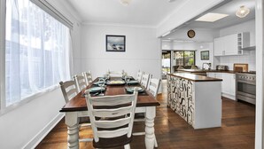 A dining area with a wooden table and white chairs, a kitchen with white cabinets and a stove, and a view of the outdoors through a window.