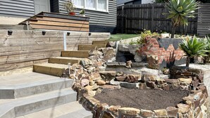 A stone fire pit with steps leading up to it, surrounded by a garden with various plants and a wooden deck.