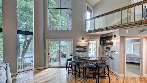 A spacious living area with a high ceiling, large windows, a kitchen with a bar, and a loft bedroom. - Lakefront w/ Private Dock + Hot Tub | Pontoon Rental | Butler Bliss by AvantStay (Sherrills Ford)