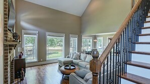 A spacious living room with a large sectional sofa, a coffee table, and a fireplace. There are large windows providing natural light and a view of the outdoors. A wooden staircase with black railings leads to an upper level. - New Dock & Custom Home | Dock Holiday by AvantStay (Huntersville)