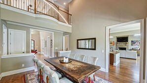 A spacious dining area with a long wooden table, cushioned chairs, and a red patterned rug. A staircase leads to an upper level. - New Dock & Custom Home | Dock Holiday by AvantStay (Huntersville)