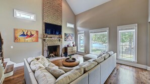 A spacious living room with a large sectional sofa, a fireplace, and large windows offering views of the outdoors. - New Dock & Custom Home | Dock Holiday by AvantStay (Huntersville)