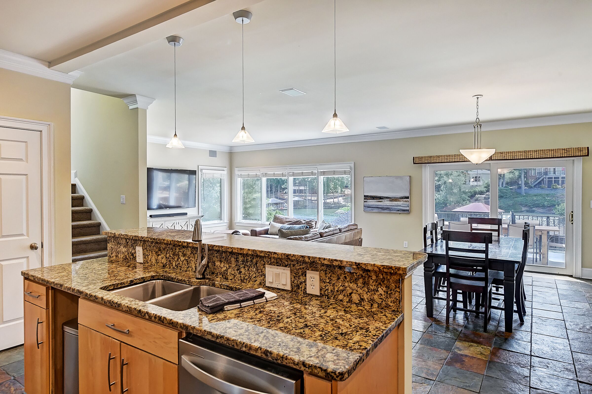 A kitchen with granite countertops, a stainless steel sink, and wooden cabinets. There is a dining area with a table and chairs, and a large window offering a view of the outdoors.