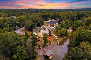 A large, two-story house with a swimming pool, surrounded by dense trees and a body of water.