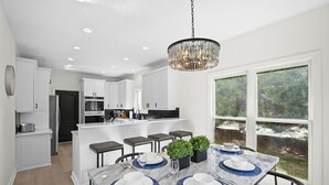 A modern kitchen with white cabinets, a marble island, and a dining area with a table set for four. - Knotty Pines by AvantStay | Lake Views, Renovated (Sherrills Ford)