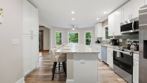 Une cuisine moderne avec des meubles blancs, un îlot central doté de tabourets de bar, des appareils en acier inoxydable et de grandes fenêtres offrant une vue sur la verdure.