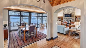 A spacious living room with a stone fireplace, large windows, and a view of a lake. - Landover Lodge by AvantStay | Waterfront + Dock (Mooresville)