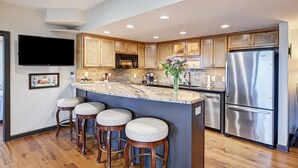 A modern kitchen with wooden cabinets, a stainless steel refrigerator, a marble countertop, and three bar stools. - Landover Lodge by AvantStay | Waterfront + Dock (Mooresville)
