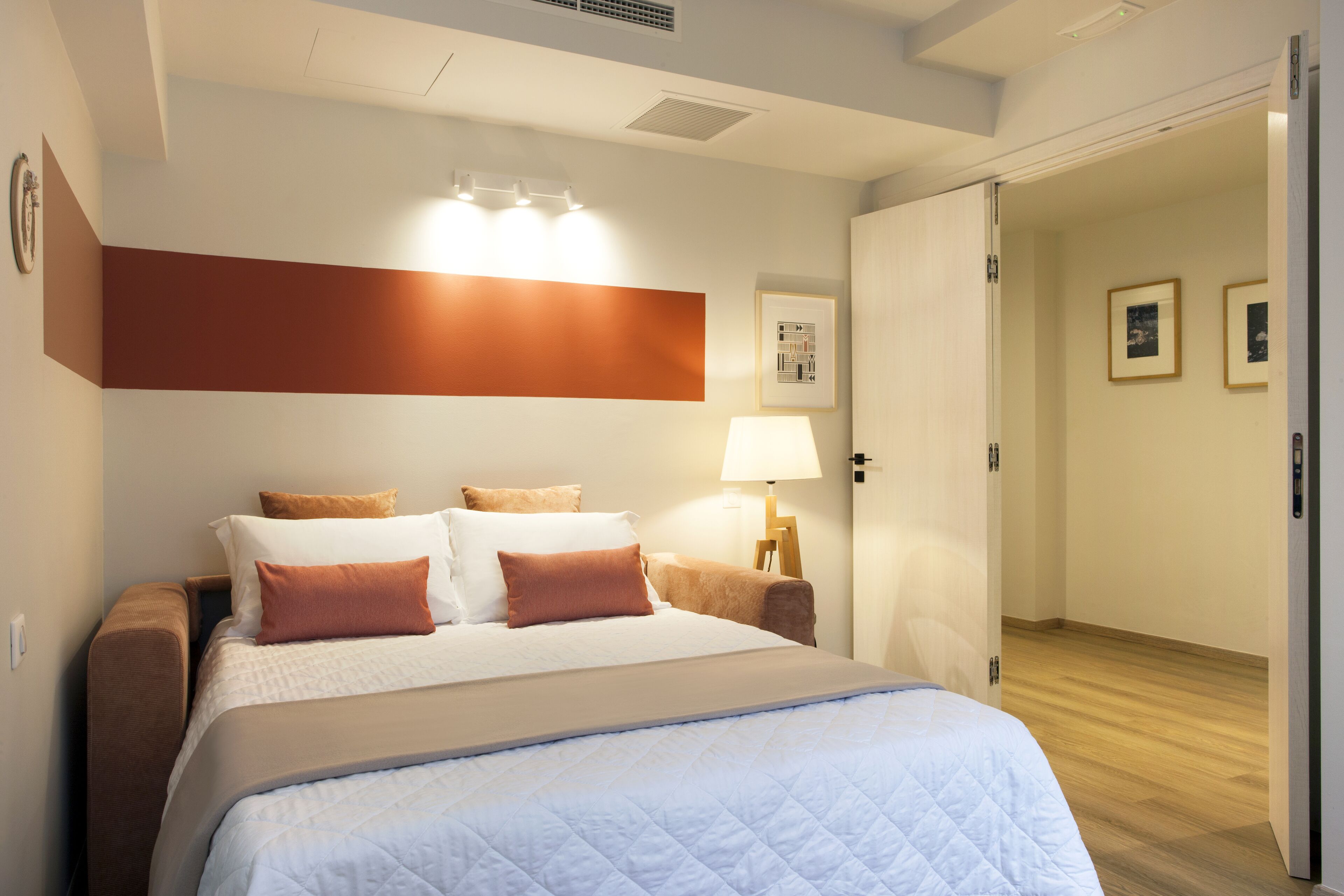 Una cama bien tendida con una colcha blanca y marrón, una lámpara de noche y una lámpara de pared.