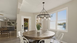 A dining area with a round wooden table, white chairs, a chandelier, and a view of a lake through large windows. - Lakefront Estate | Pool + Hot Tub + Dock + Sauna | Oasis at Ballycastle (Mooresville)