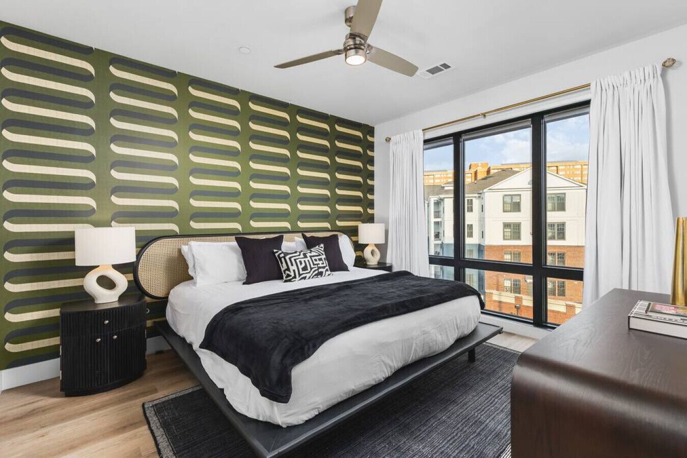 Un dormitorio moderno con una cama grande, un ventilador de techo, vistas de edificios a través de un ventanal amplio y una pared con diseño.