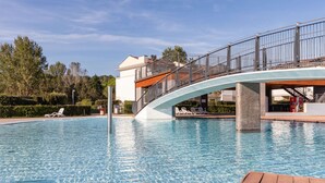 Zwembad met houten terras op de voorgrond, een gebogen brug met leuningen, en een gebouw met een witte gevel achterin.