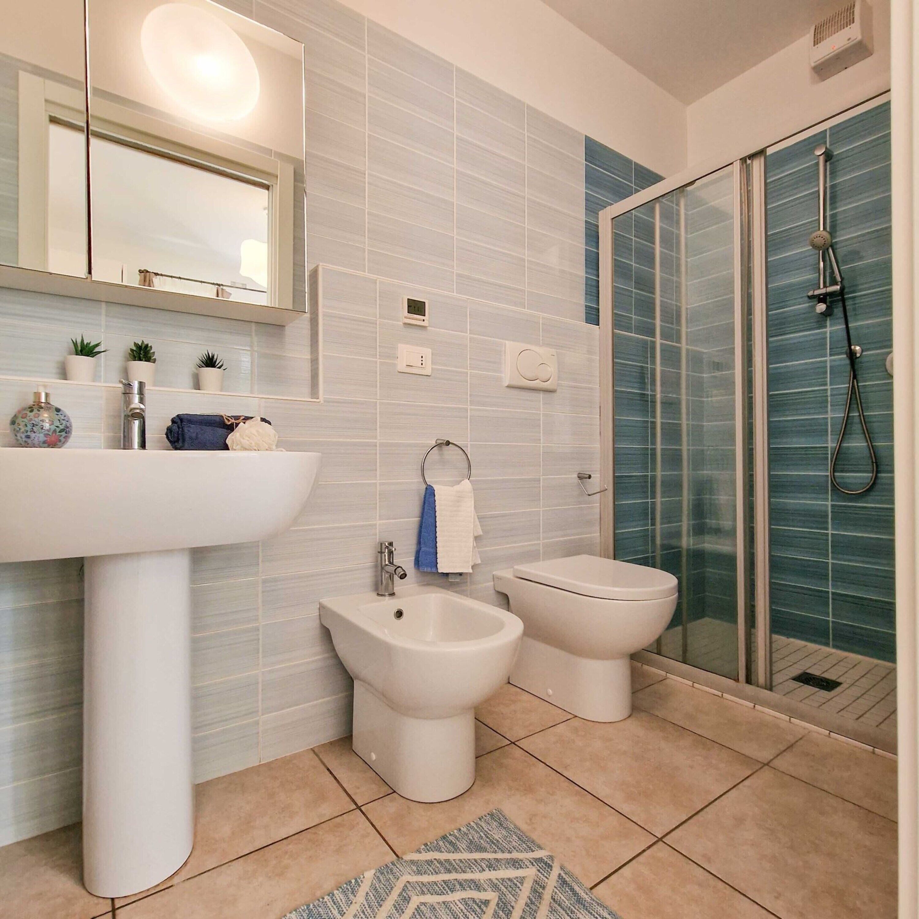 A modern bathroom with a white sink, bidet, and toilet, a glass shower enclosure, and a mirror with a light above it.