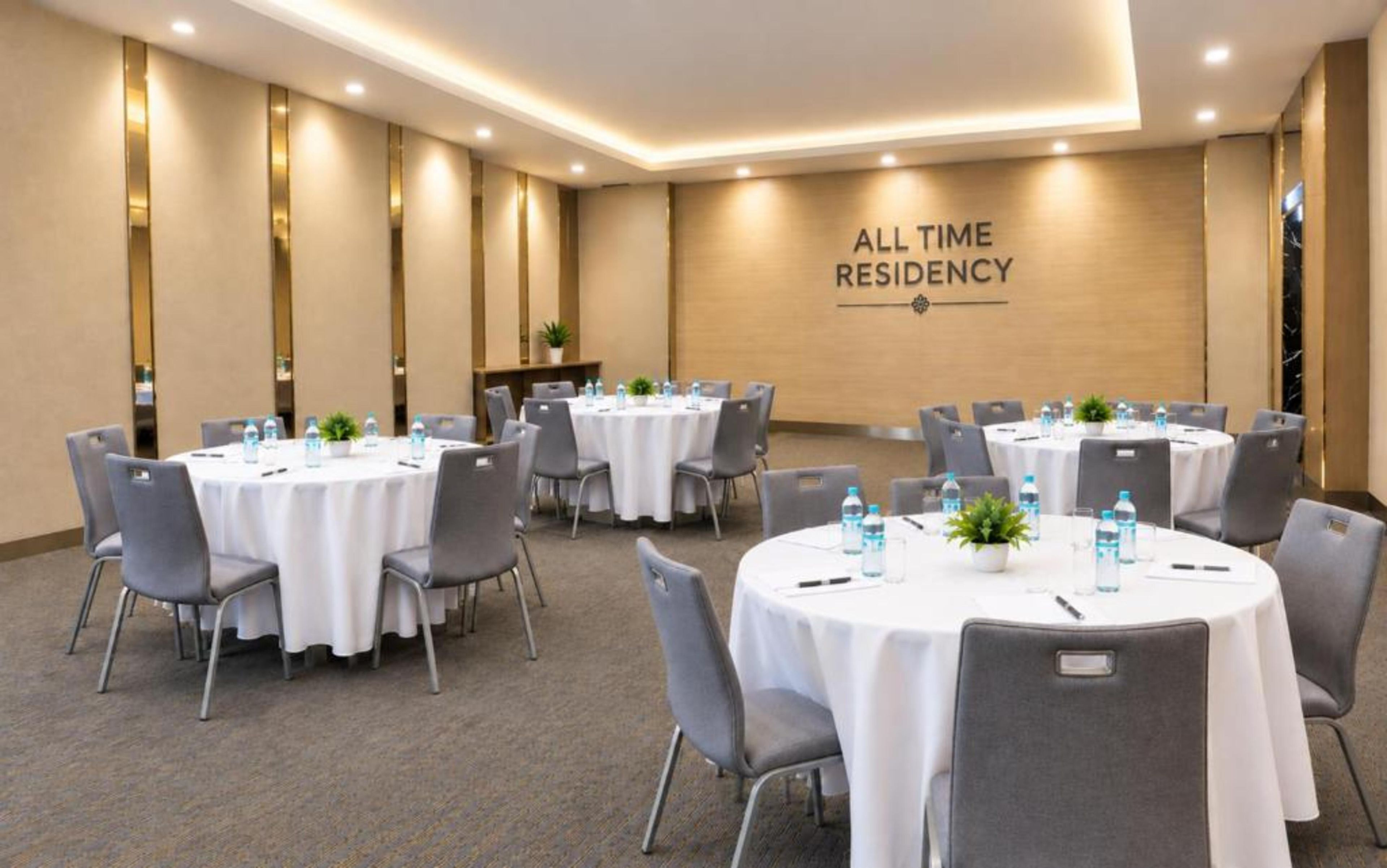 Un salón de conferencias con varias mesas redondas, cada una con sillas y botellas de agua, en un hotel llamado 'ALL TIME RESIDENCY'.
