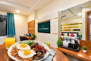 A hotel room with a dining area featuring breakfast, a TV screen with 'All Time Residency' logo, and a bed with pillows and a bedside table.
