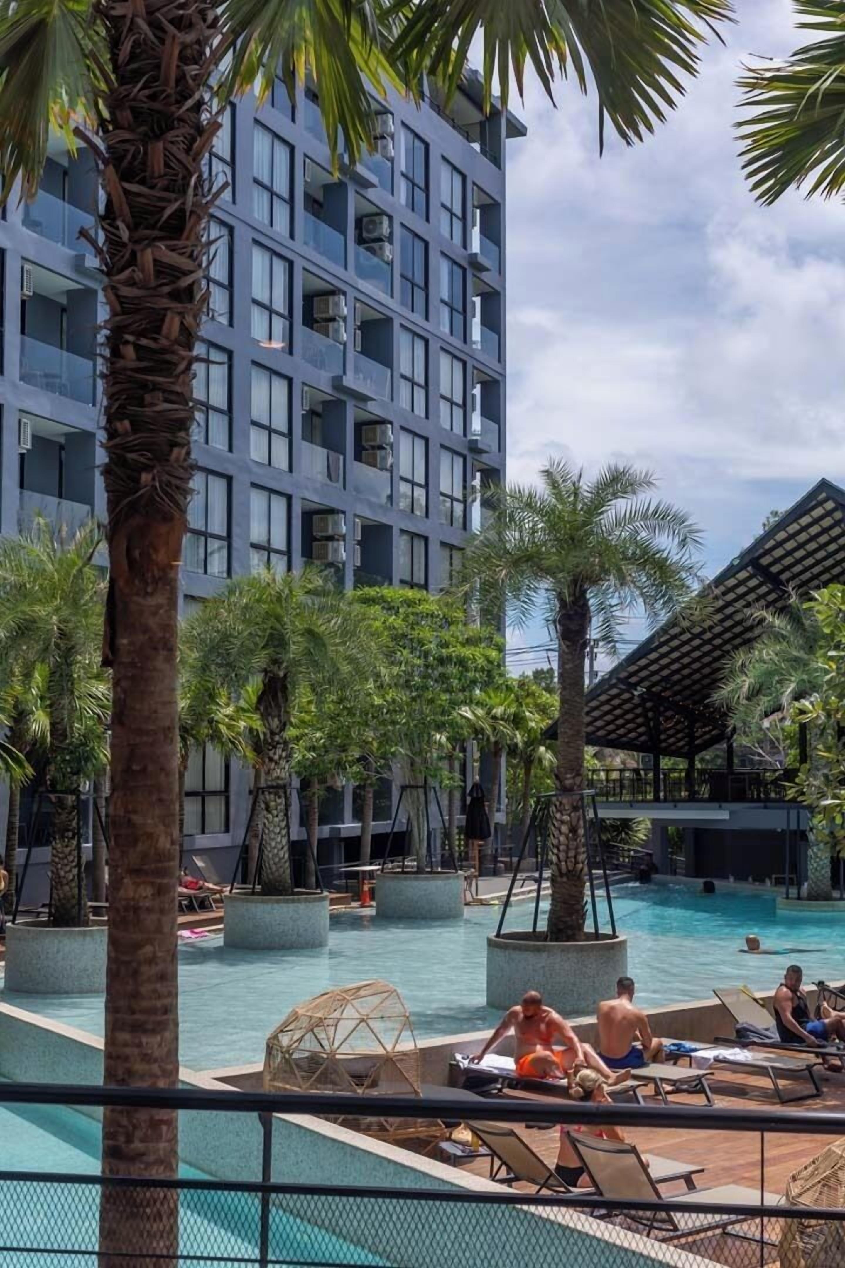 A modern hotel with a swimming pool, palm trees, and lounge areas.