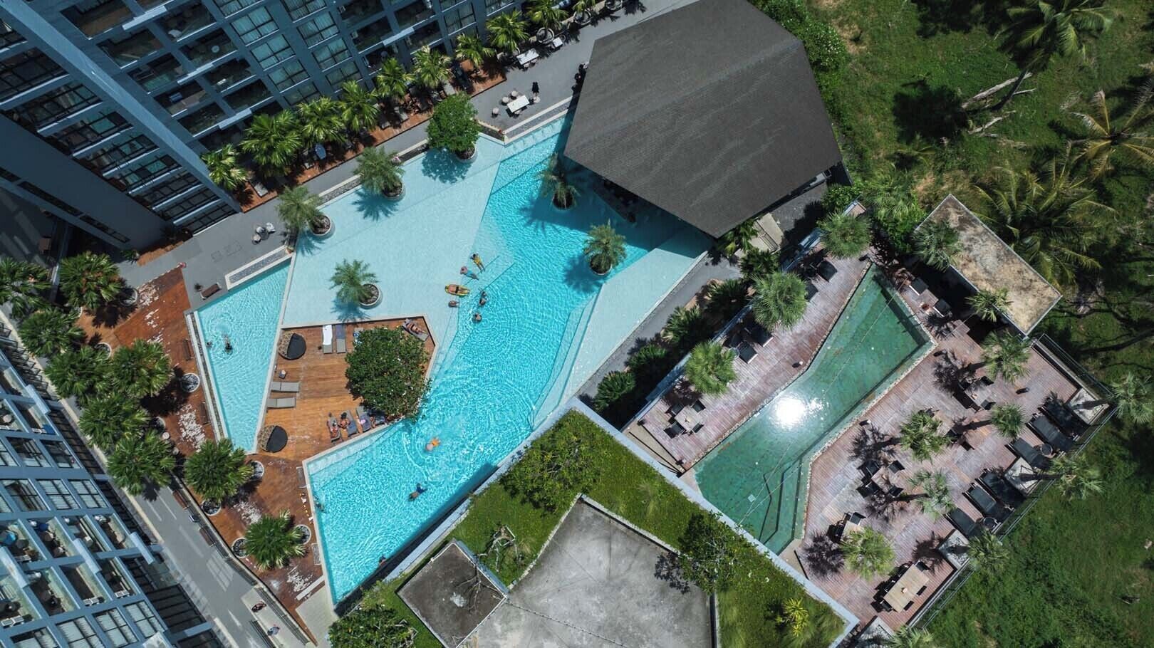 An aerial view of a hotel with a large central swimming pool surrounded by palm trees and lounge areas.