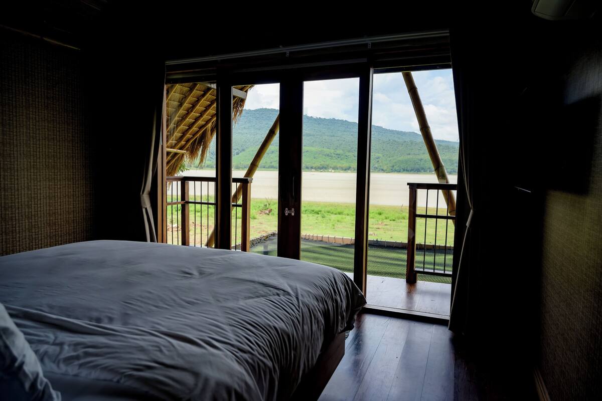 A bedroom with a large bed and a view of a river and mountains through open doors.