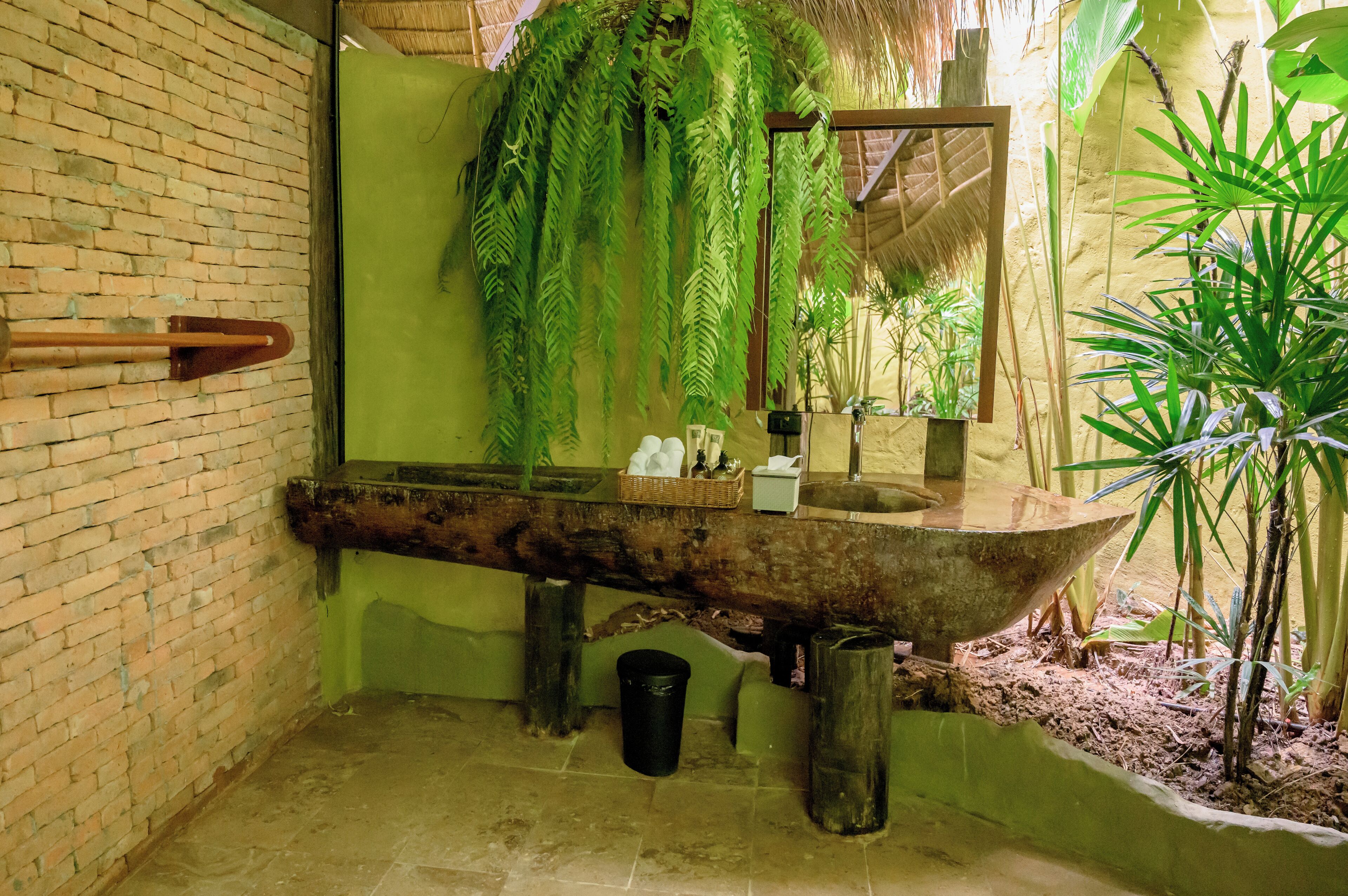 A rustic bathroom with a brick wall, a wooden basin, hanging ferns, and a large mirror.