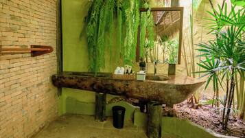 A rustic bathroom with a brick wall, a wooden basin, hanging ferns, and a large mirror.