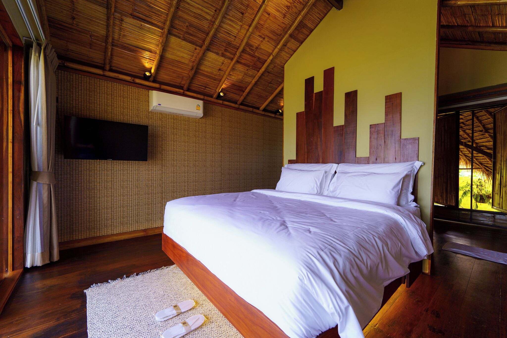 A bedroom with a wooden ceiling, a flat-screen TV mounted on the wall, a bed with white bedding, and a large window with a view of greenery.