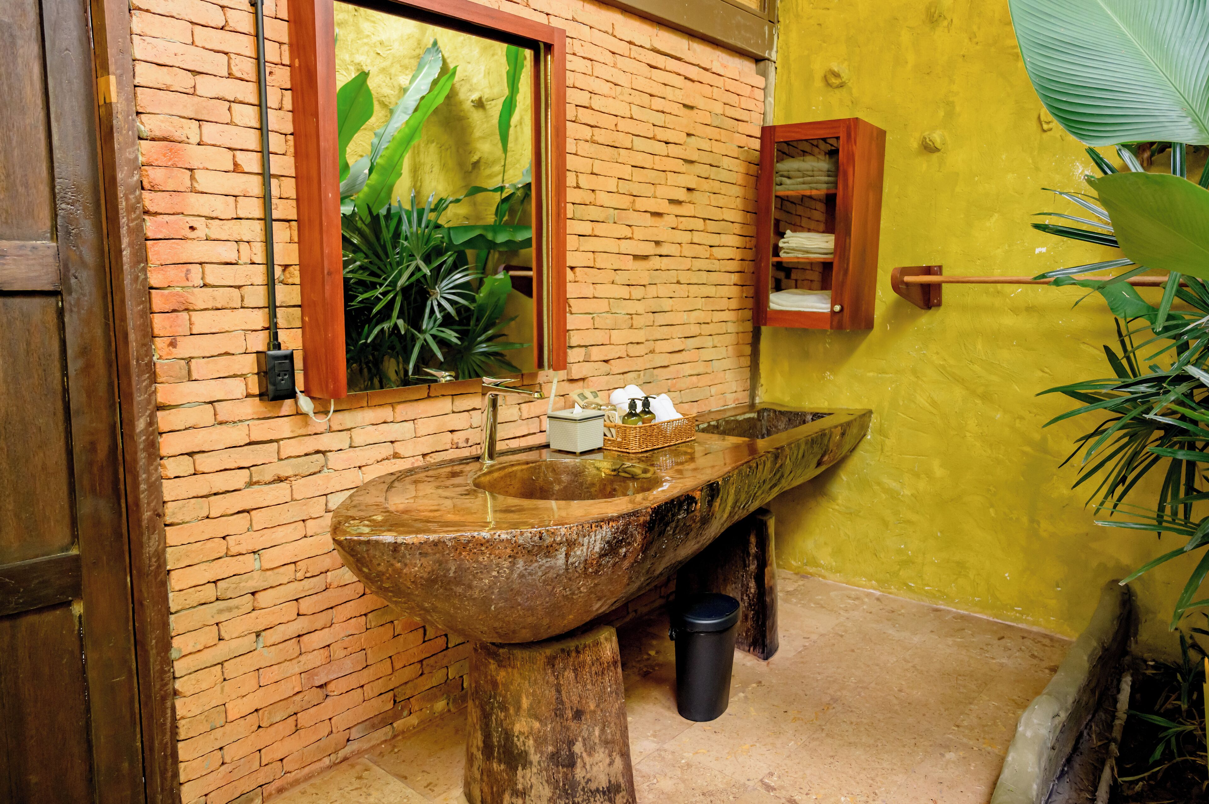 A rustic bathroom with a large stone sink, a wooden door, and a mirror with a view of green plants.
