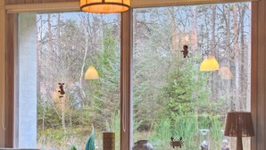 A cozy room with a large window overlooking a forest. There are two hanging lamps, a table with a kettle, and various decorative items.