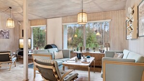A spacious living area with a wooden ceiling, large windows offering a forest view, a fireplace, and modern furniture.