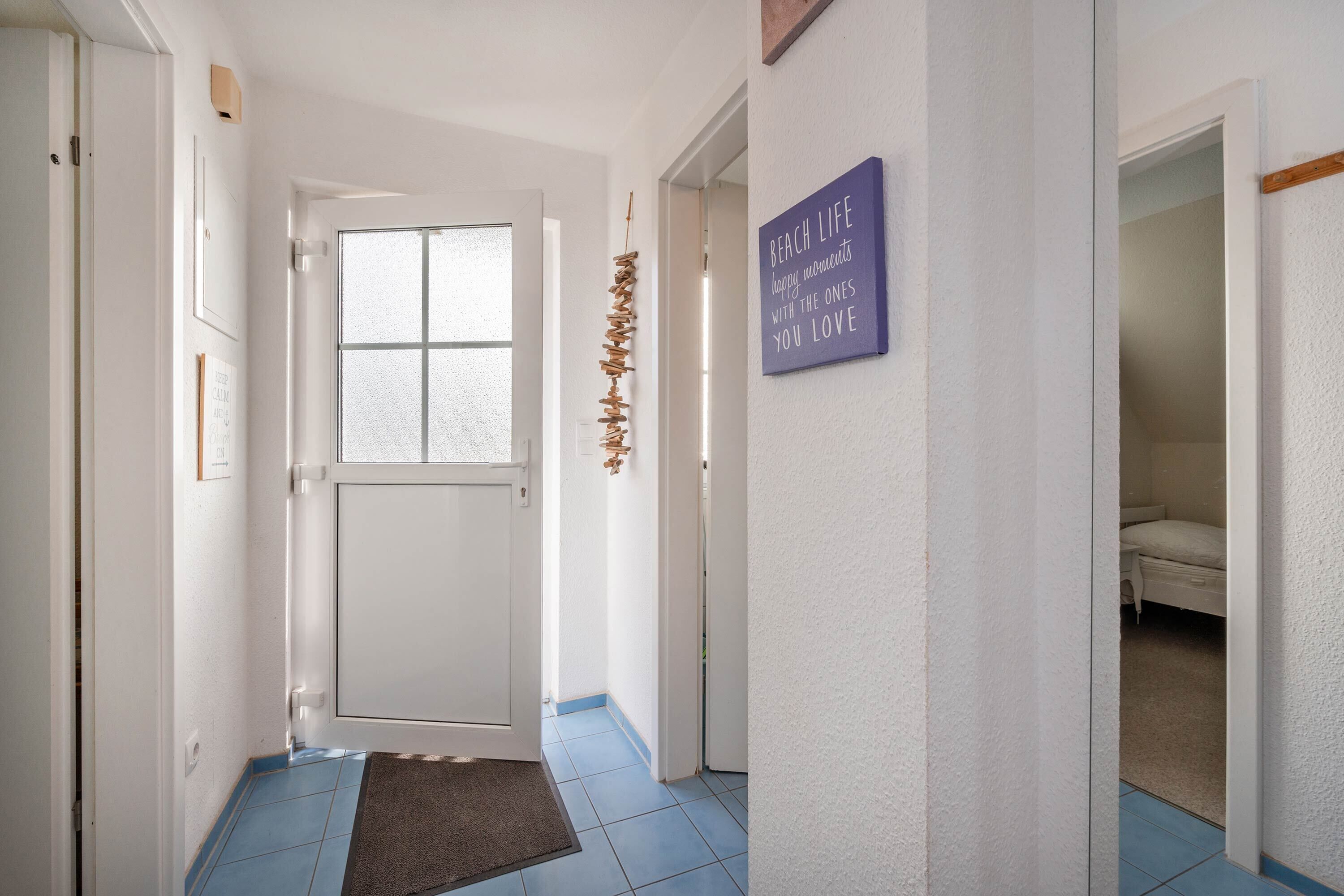 Een gang met een witte deur, een vloer betegeld met blauwe tegels, een deurmat en een bordje aan de muur.