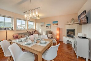 A modern dining area with a wooden table set for four, white chairs, a chandelier, and a fireplace with a mounted television. - Ferienwohnung Strandbrise (Koserow)