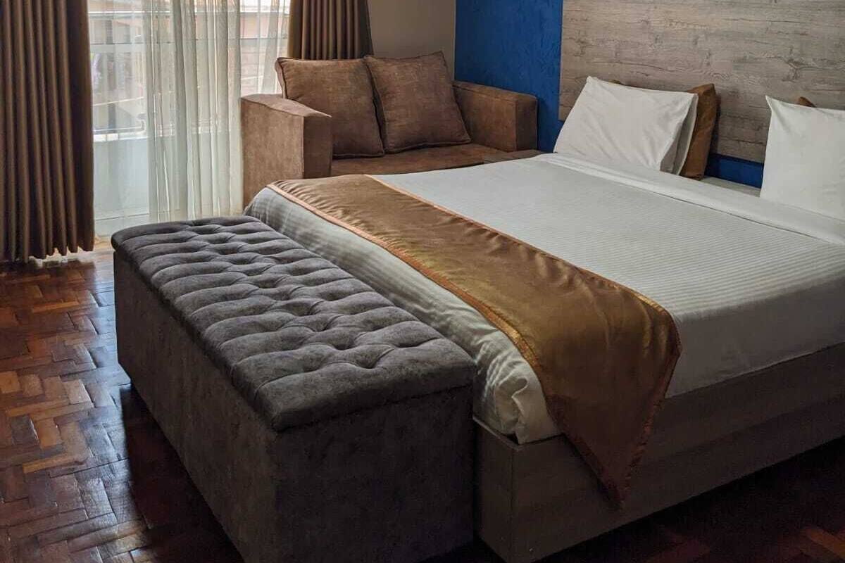 A modern hotel room with a large bed, a bench, and a view of the outside through sheer curtains.