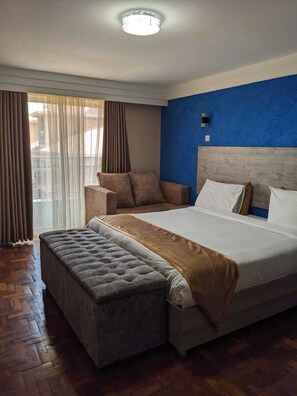A modern hotel room with a large bed, a bench, and a view of the outside through sheer curtains. - Essence Hotel (Ndenderu)