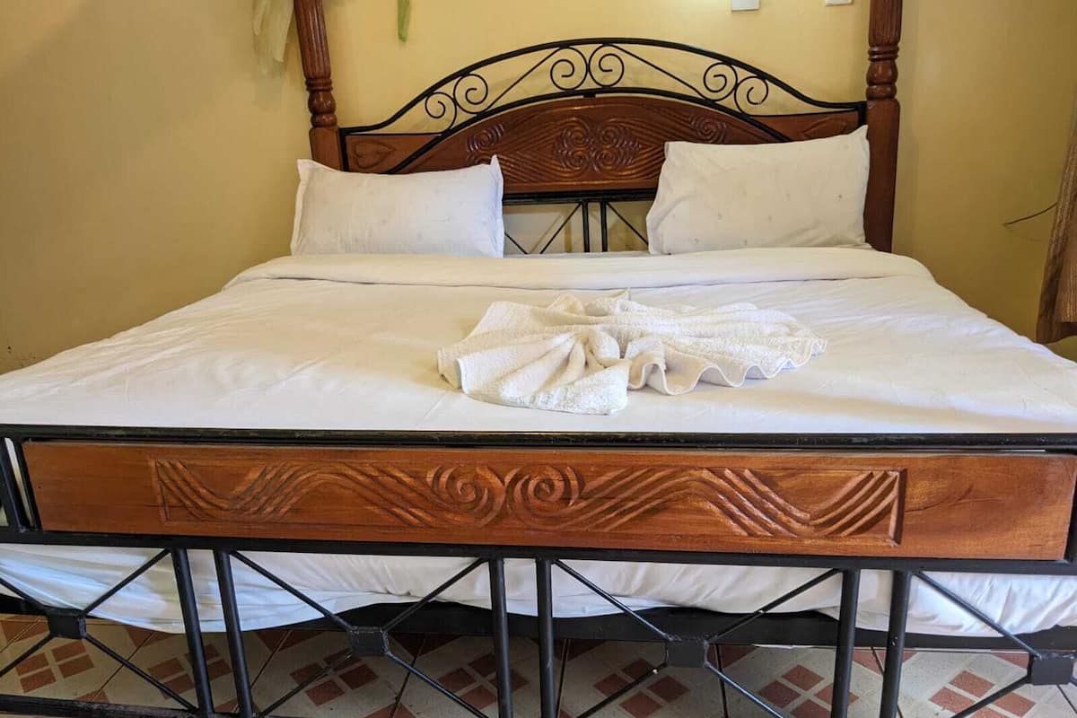 A neatly made bed with white linens and a folded towel, a wooden headboard with intricate carvings, and a tiled floor with a geometric pattern.