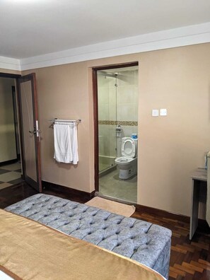 A hotel room with a bed, a bathroom with a toilet and shower, and a wooden floor. - Essence Hotel (Ndenderu)
