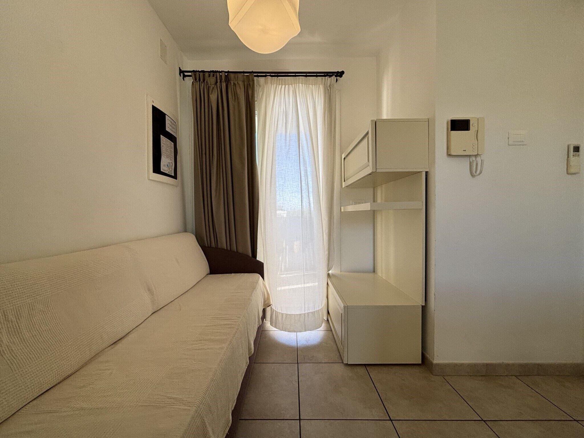 A room with a beige sofa, a small table, a white shelf unit, a telephone, and a window with curtains.