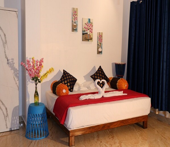 A neatly made bed with a red bedspread and decorative pillows, a blue stool with a vase of flowers, and three framed artworks on the wall. - Thaalam-an ayurvedic Resort (Yercaud)
