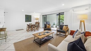 A modern living room with a sofa, coffee table, and chairs. There is a dining area with a table and chairs. A large window provides natural light and a view of greenery outside. - Lindy Lou's Entire Rental Unit in Sawtell (SAWTELL)