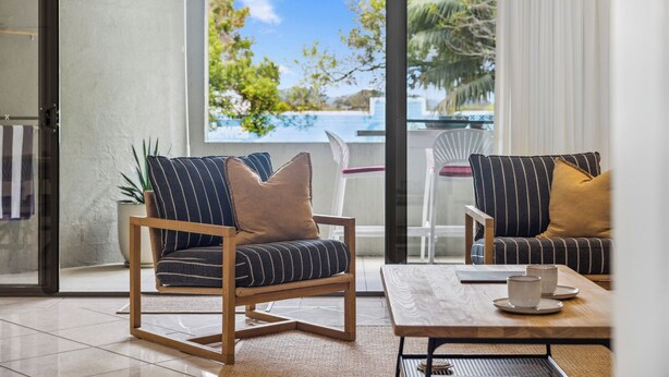 A modern living room with a glass door leading to a balcony, featuring striped sofas, a wooden coffee table, and a view of a pool and trees. - Lindy Lou's Entire Rental Unit in Sawtell (SAWTELL)
