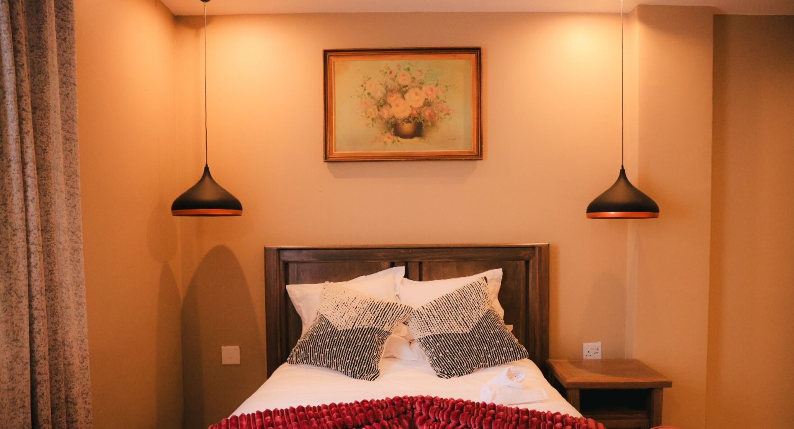 A neatly made bed with a red blanket, two black lamps, and a painting on the wall.