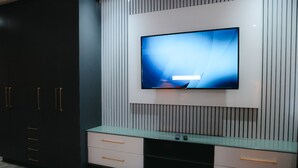 Une chambre moderne dotée d’un grand téléviseur à écran plat fixé au mur, lequel est orné de lignes verticales, d’une armoire sombre avec de nombreux tiroirs et d’une petite table en verre sur laquelle reposent deux télécommandes.